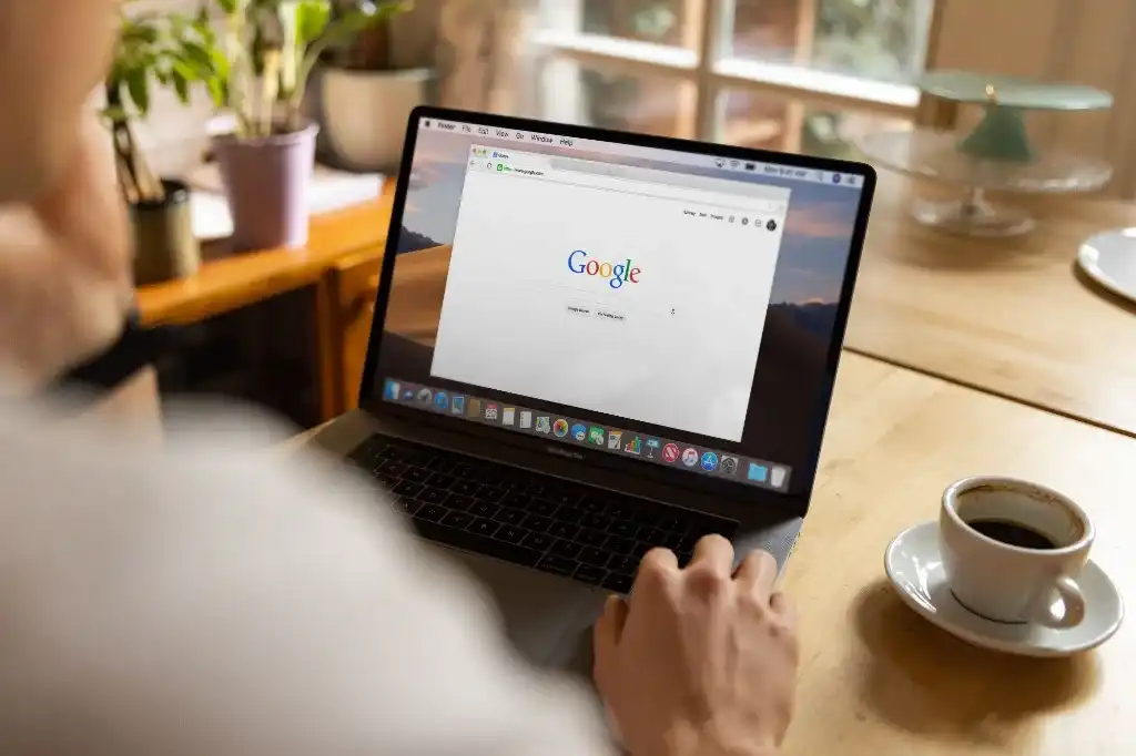 Pessoa usando um MacBook com a página inicial do Google aberta, ao lado de uma xícara de café